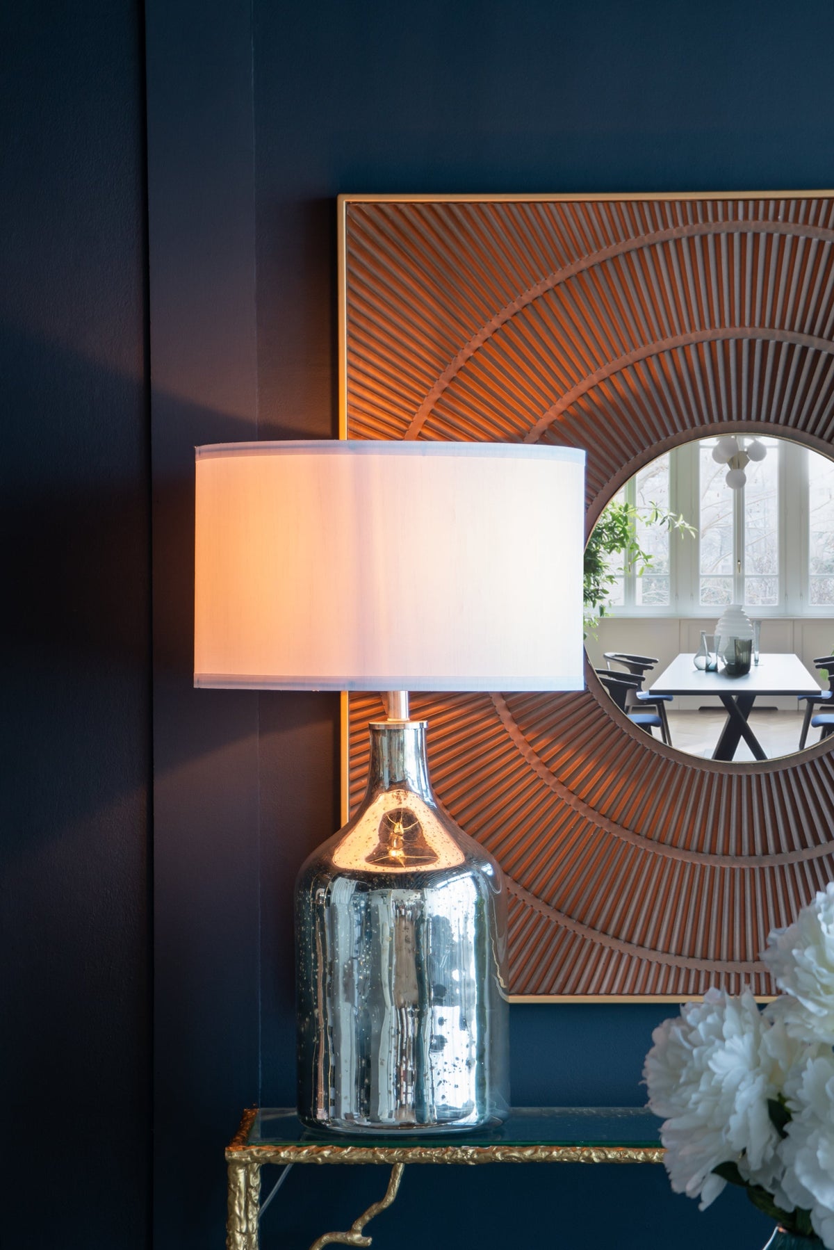 Silver round table lamp with white shade and reflective base beside mirror on dark blue wall on Tampa Home & Garden Store