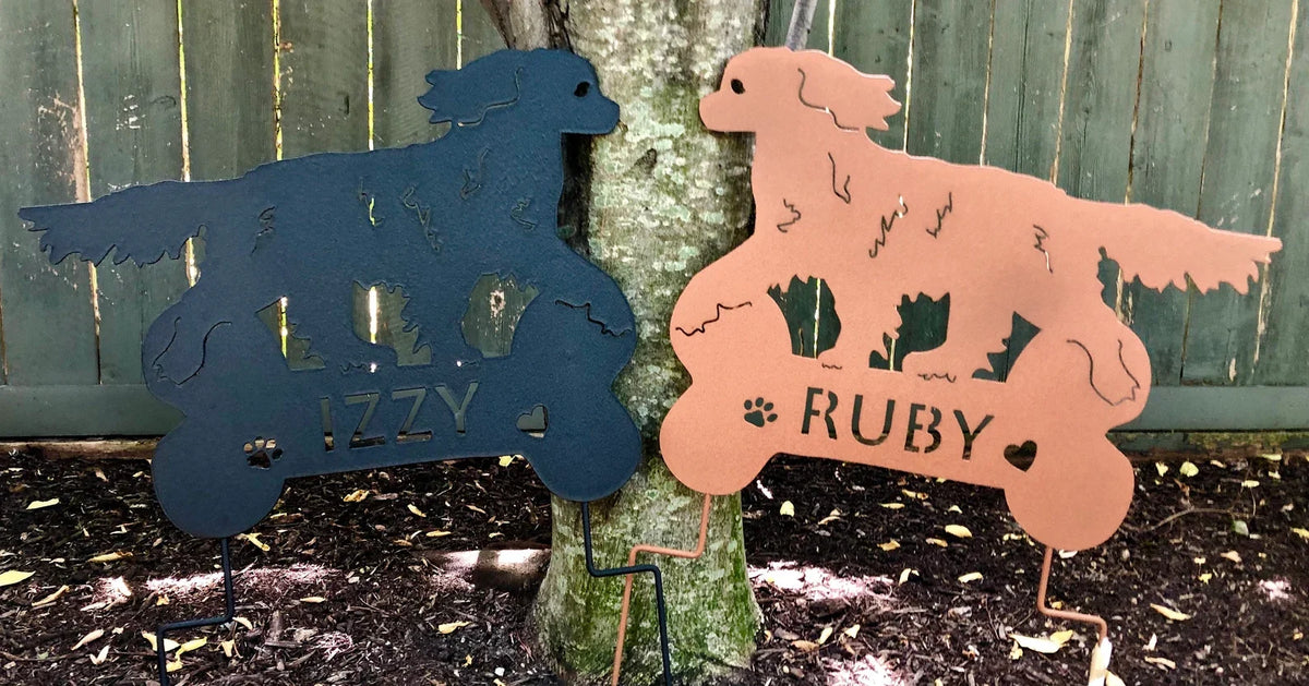 Two King Charles dog sculptures in blue and pink metal yard art by Rustica Ornamentals featuring IZZY and RUBY names on Tampa Home & Garden Store