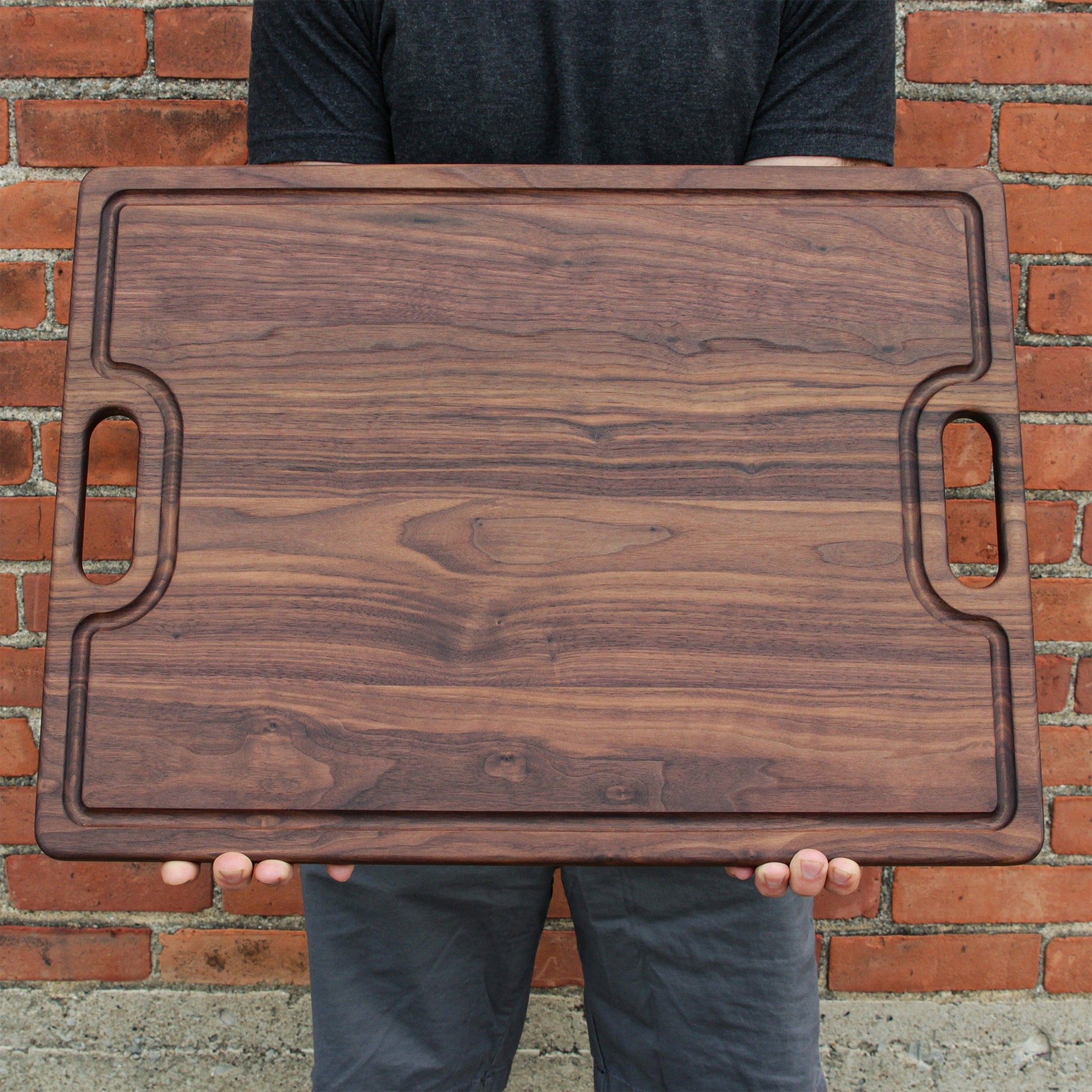 Dark walnut cutting board with built-in handle cutouts, held against a brick wall on Tampa Home & Garden Store