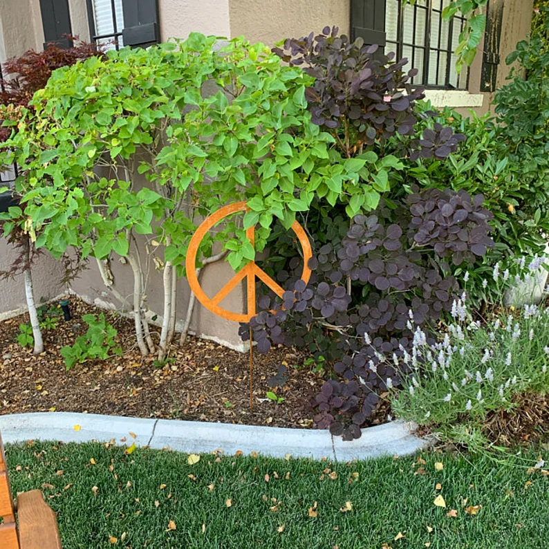 Outdoor peace sign garden stake, orange metal yard art among green and purple foliage in a yard on Tampa Home & Garden Store
