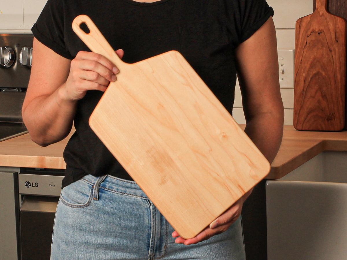 Wide paddle board cutting board in light wood held by a person in a black shirt in a kitchen on Tampa Home & Garden Store