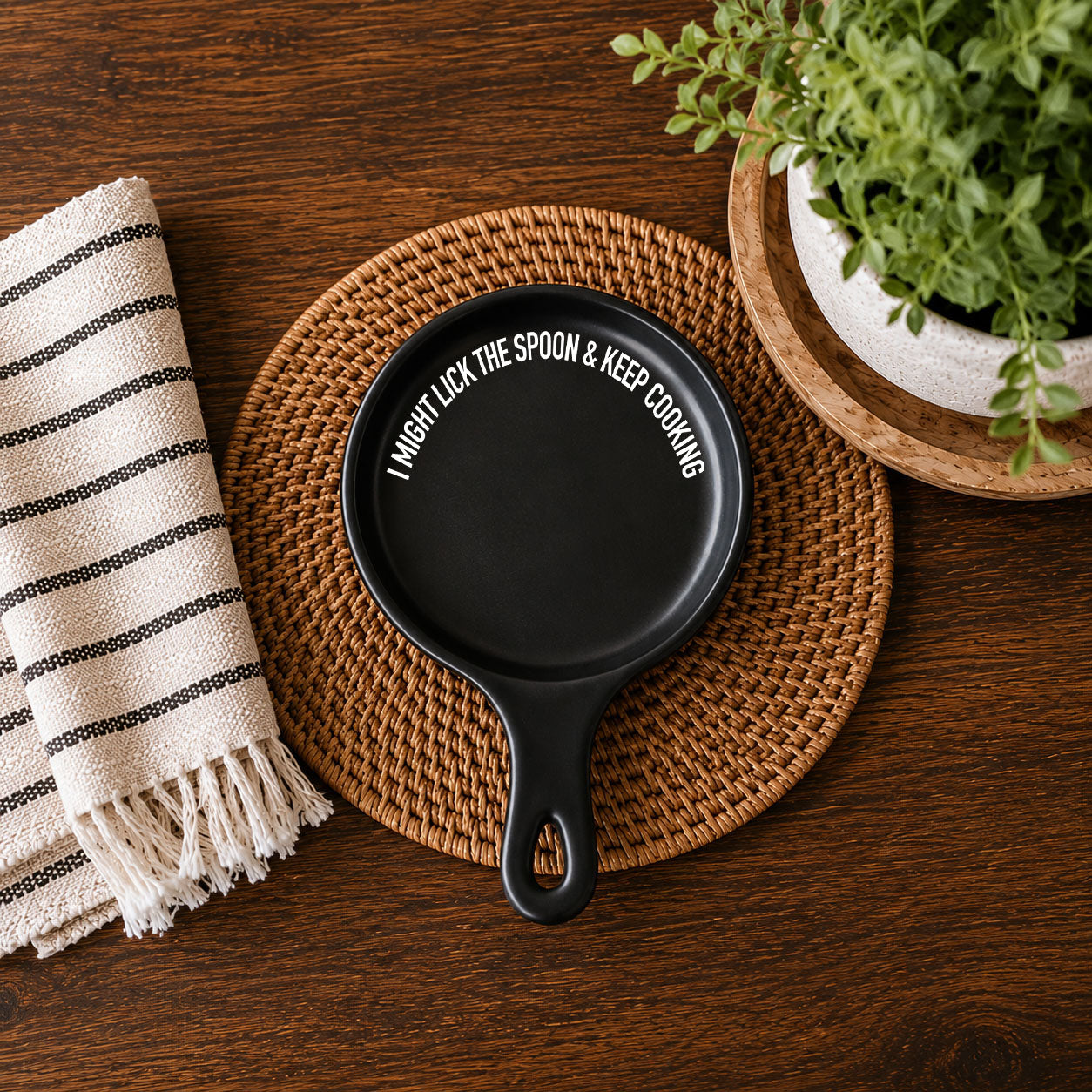 Gia Roma black spoon rest with curved white slogan I might lick the spoon & keep cooking on woven placemat beside striped napkin and plant on Tampa Home & Garden Store