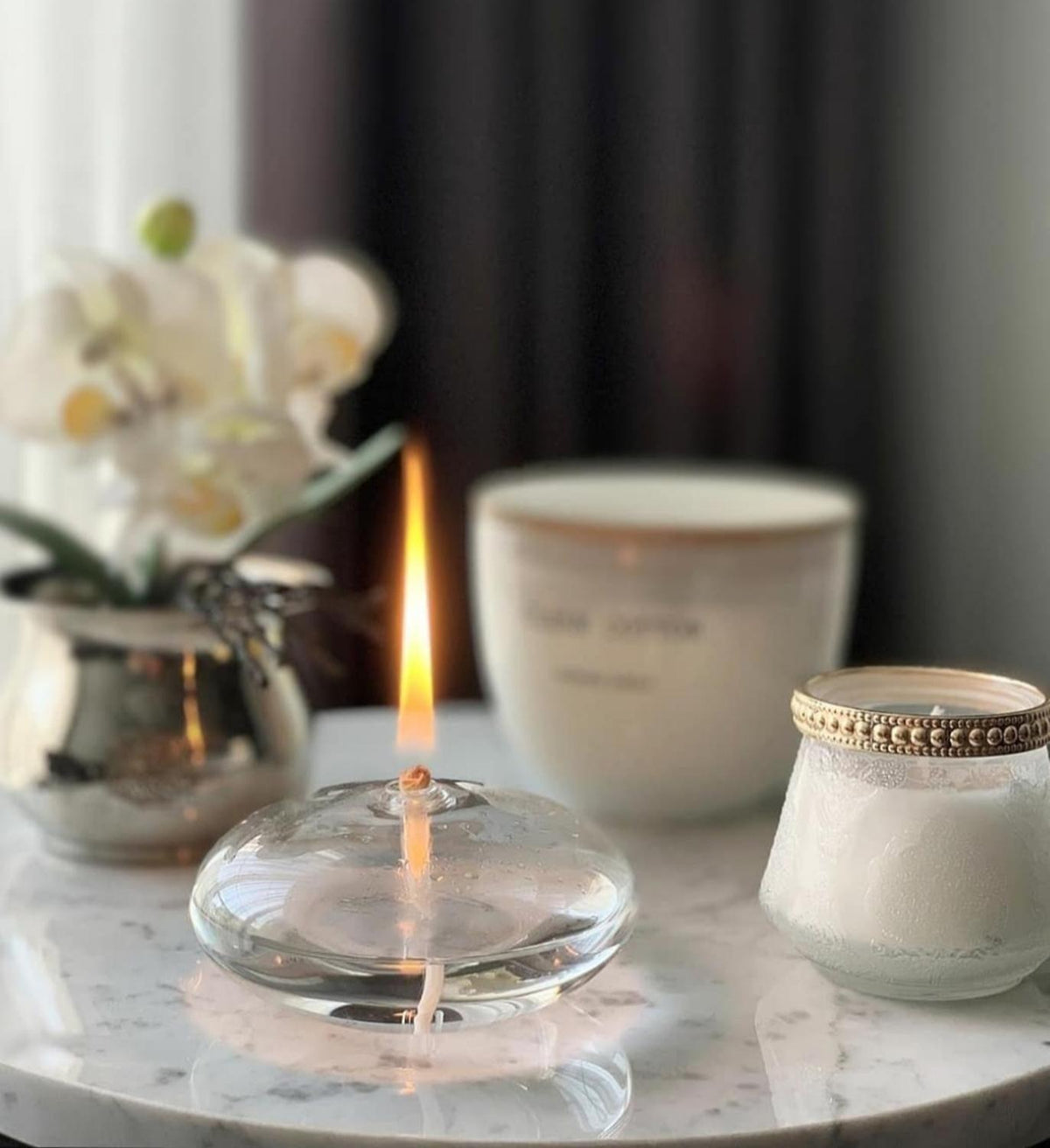 Round glass oil candle with burning wick on marble table beside a white decorative vase on Tampa Home & Garden Store
