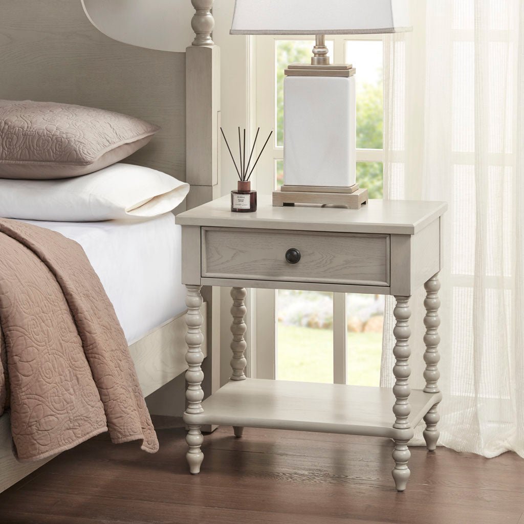 Nightstand with 1 drawer and turned legs in light gray wood finish beside a bed on Tampa Home & Garden Store