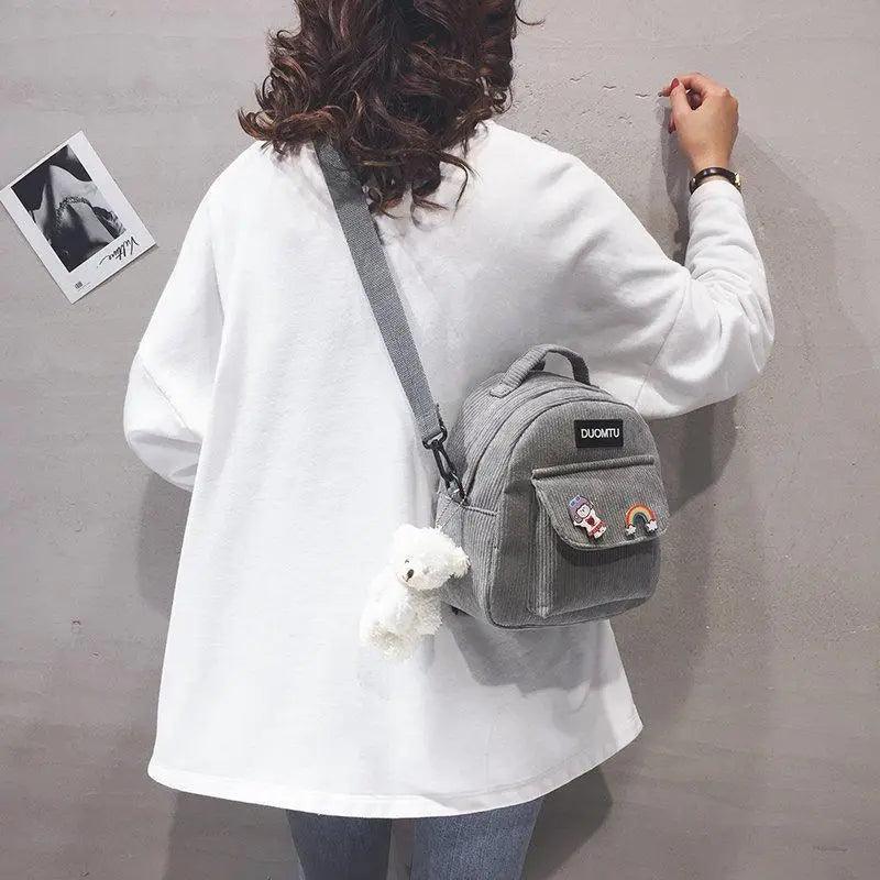Stevvex small gray backpack with front pocket patches and a white teddy keychain worn crossbody on Tampa Home & Garden Store